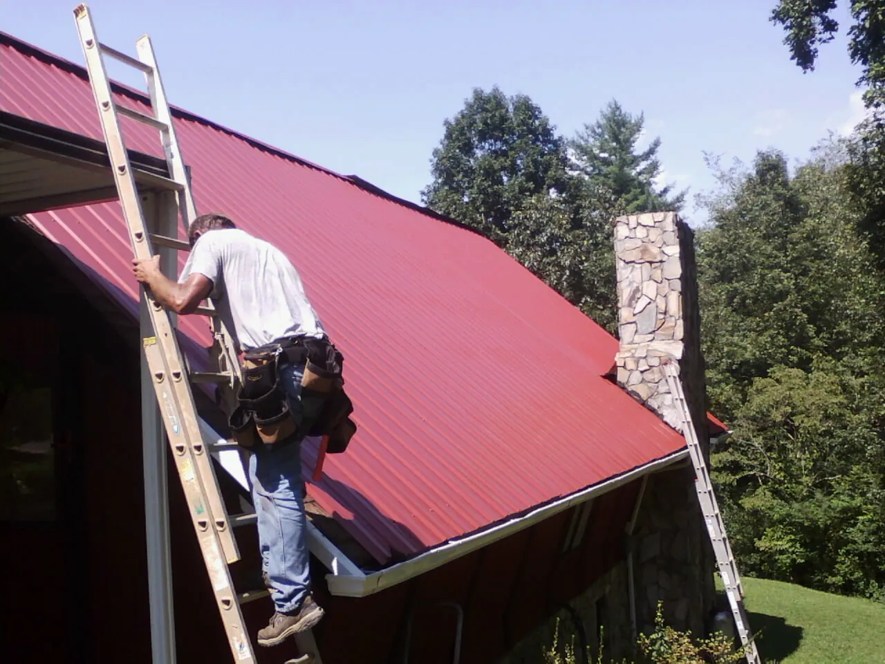 Roofer on a ladder beside a red metal roof and stone chimney — Soffit & Fascia Repair by Roofing Brothers in Shedd